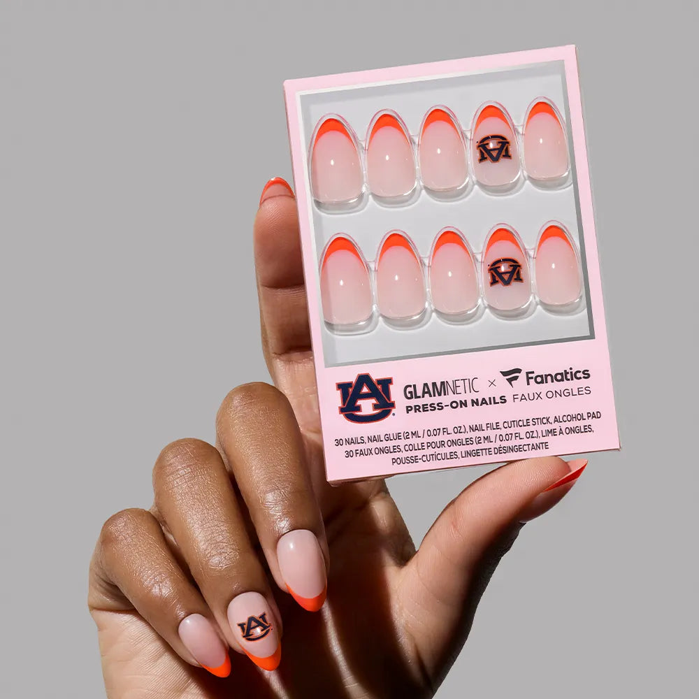 A hand holds a glamnetic Auburn Tigers Press-On Nail Set box, with orange tips and team logo nail art. One nail with the Auburn logo is worn on their finger.