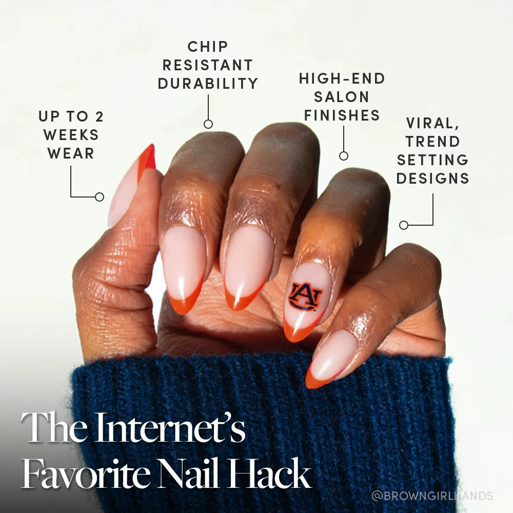 A hand with long, almond-shaped nails features the Auburn Tigers Press-On Nail Set by glamnetic, showcasing a French tip and "AU" logo. Enjoy up to 2 weeks of chip-resistant wear, high-end finishes, and viral trendsetting designs.