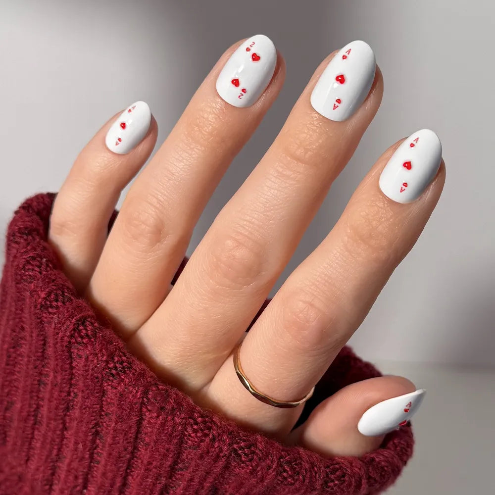 A hand wears glamnetic's Ace of Hearts short oval press-on nails—white with small red hearts and a tiny red letter, resembling ace of hearts designs. The person accessorizes with a gold ring and a burgundy knit sweater.