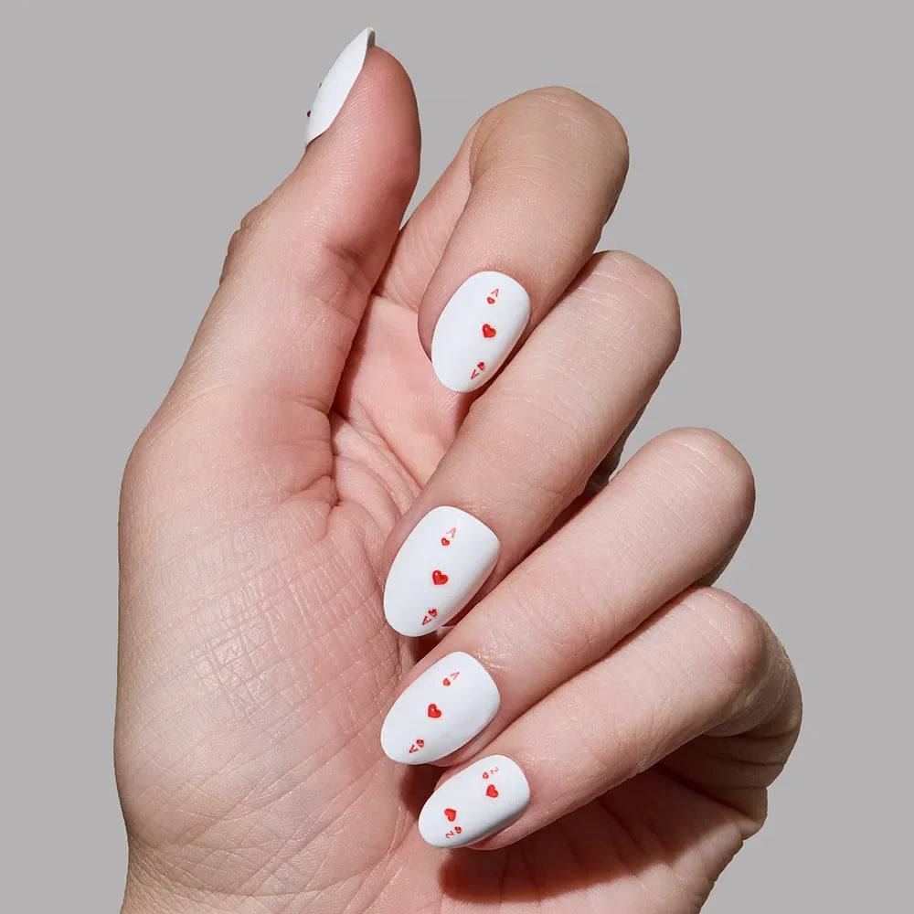 A hand displays glamnetic's Ace of Hearts press-on nails—long, oval, white nails adorned with small red heart designs—against a plain gray background.