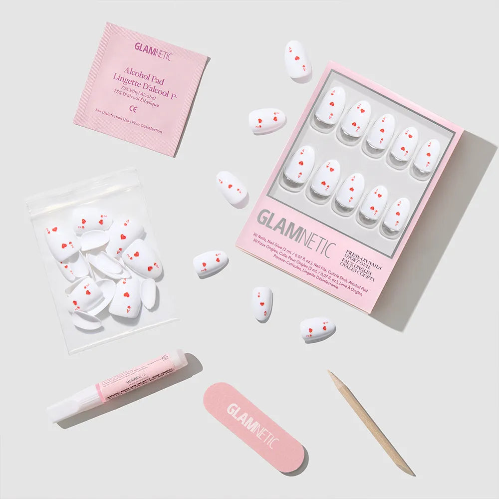 The Ace of Hearts by glamnetic is a white oval press-on nail set with small red hearts, including a pink file, wooden cuticle stick, nail glue, alcohol pad, and extra nails—all neatly displayed on a light gray surface.