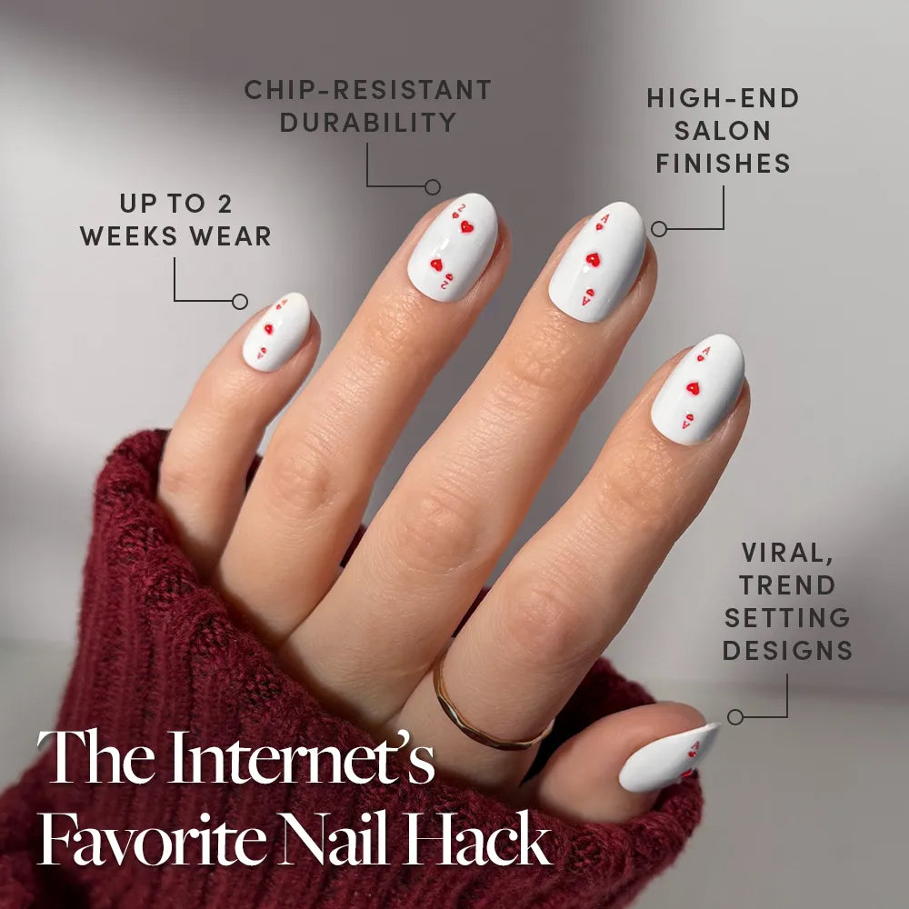 A hand wears glamnetic's Ace of Hearts oval press-on nails—white with small red hearts—and a burgundy sweater. Features include chip resistance, luxe finishes, up to 2 weeks’ wear, and trendsetting style.