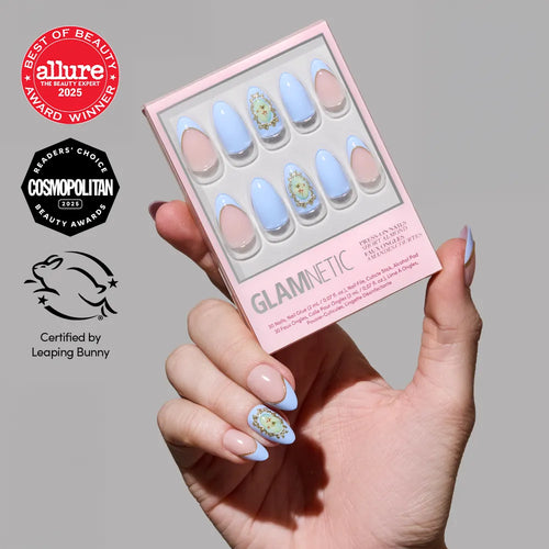 A hand with pastel blue and nude floral short almond nails holds a box of glamnetic Angel in Blue nails. Allure, Cosmopolitan beauty awards, and Leaping Bunny certification logos appear on a plain gray background.