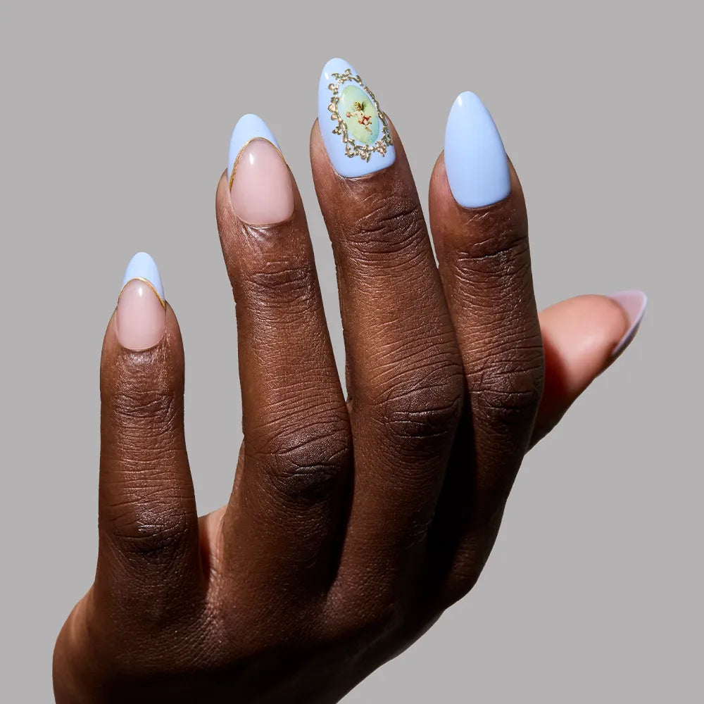 A hand with short almond nails features glamnetic’s Angel in Blue: three nails in light blue (one with a floral design), two in natural nude, and elegant 3D gold accents, all set against a plain gray background.