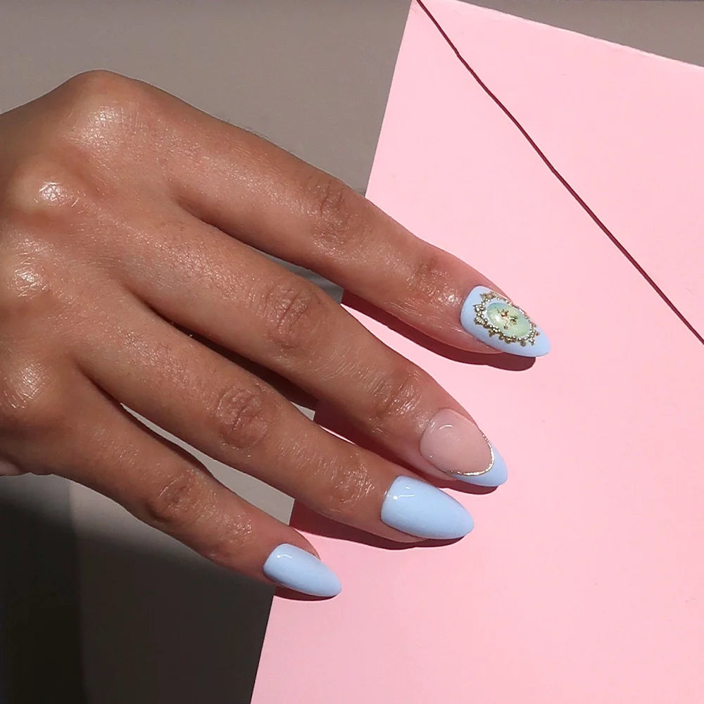 A hand with short almond nails painted in Angel in Blue by glamnetic, featuring light blue and nude tones with a 3D gold gemstone design on the ring finger, rests on a pink envelope.