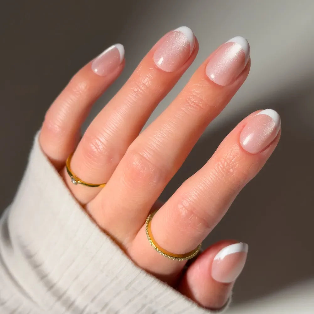 A hand with a light skin tone, wearing two gold rings and a cream ribbed sweater, displays glamnetic's Angelic Touch: short oval pink nude velvet nails with crisp white French tips.