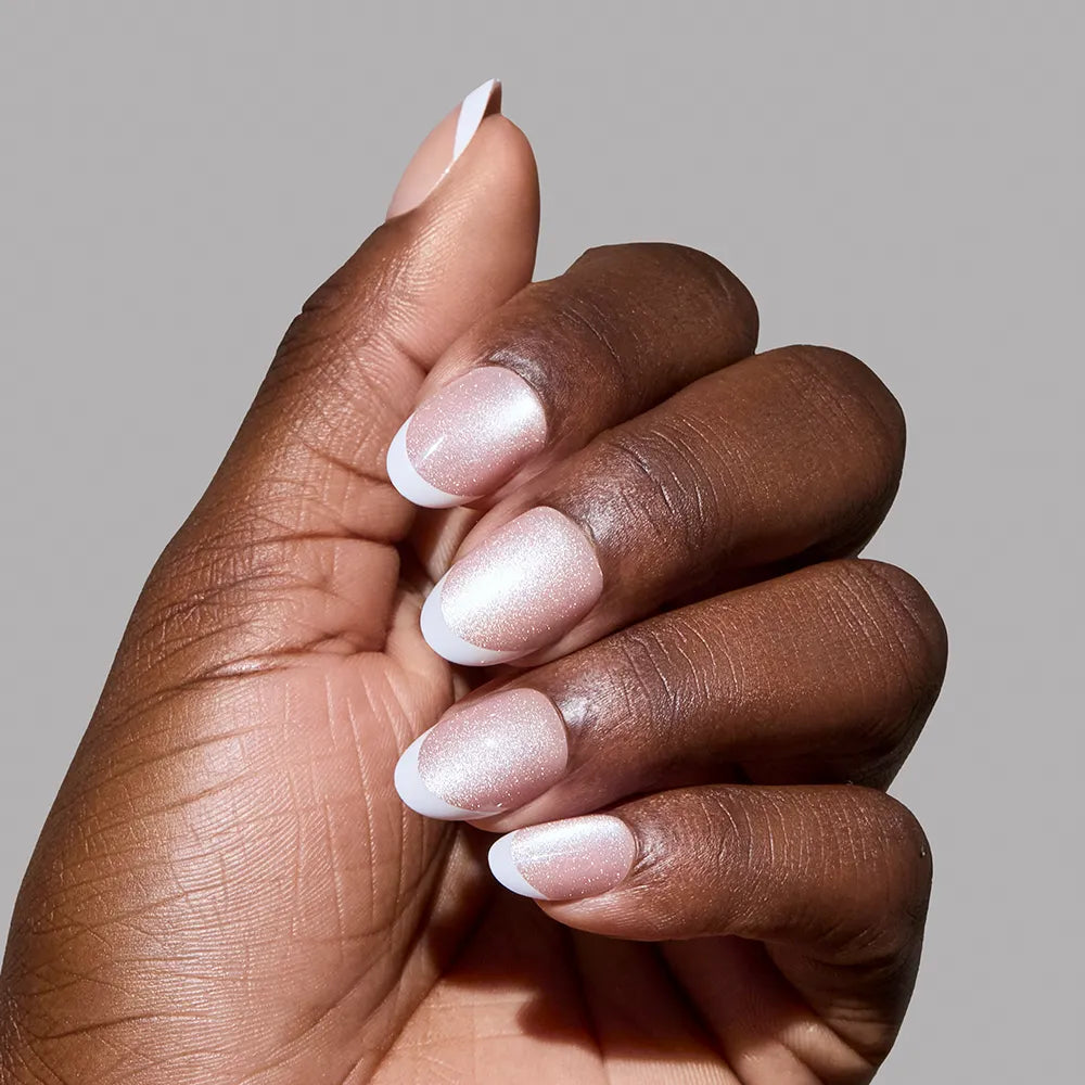 A hand with medium brown skin displays glamnetic's Angelic Touch: short oval nails in a nude velvet style with soft pink bases and crisp white French tips, set against a plain gray background.