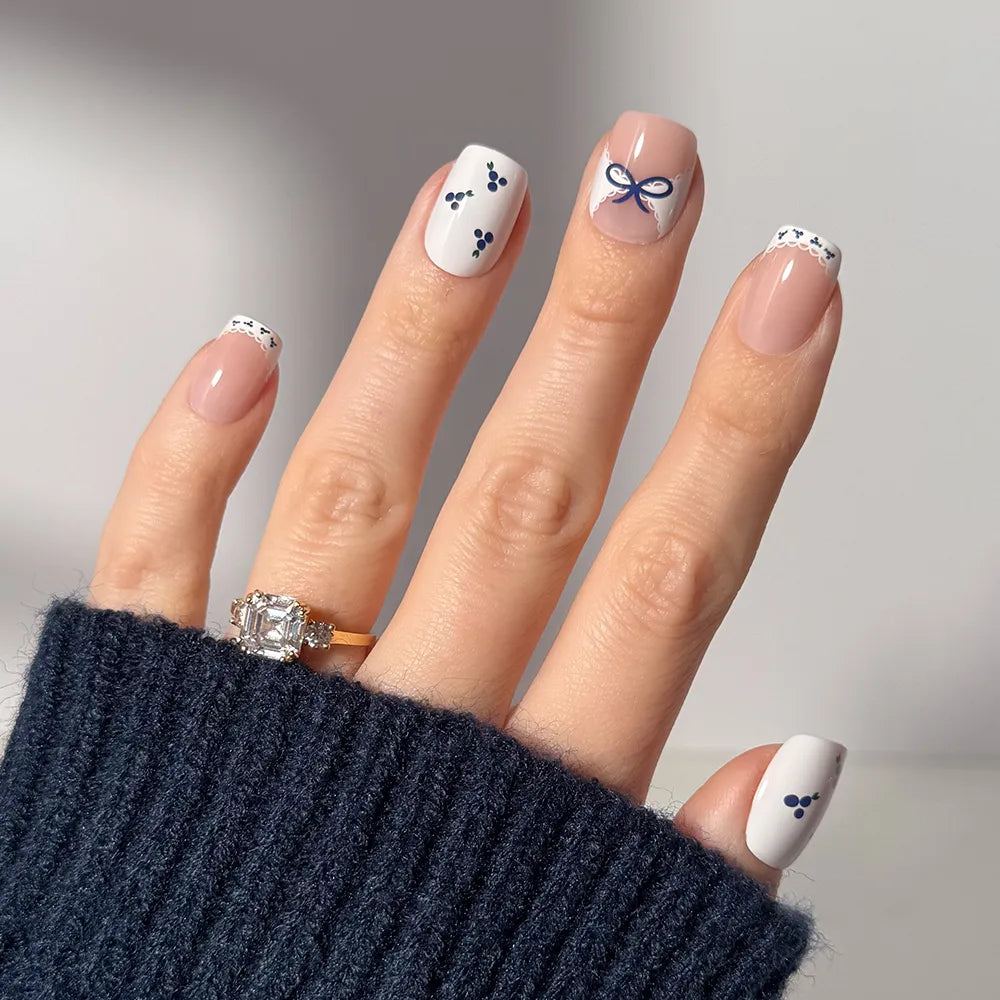 A hand with a large square-cut diamond ring displays glamnetic's Blue A Kiss squoval press-on nails, featuring a white and navy blue design with bows, dots, French tips, and blueberry accents. The person is wearing a dark knit sweater.