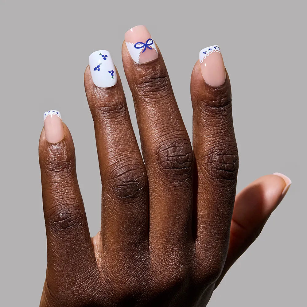 A manicured hand with glamnetic’s Blue A Kiss press on nails—squoval, nude base, and blueberry accents (dots, florals, bow on the middle finger)—set against a gray background. Perfect for anyone seeking unique nail charm.