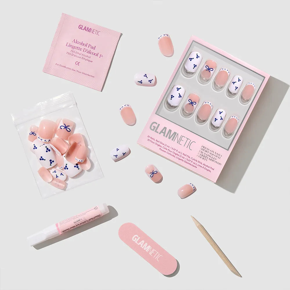 Blue A Kiss by glamnetic: squoval press-on nails with pink and blue bow designs and blueberry accents. Includes nail glue, alcohol pad, emery board, wooden cuticle stick, and resealable pouch, all displayed on a light surface.
