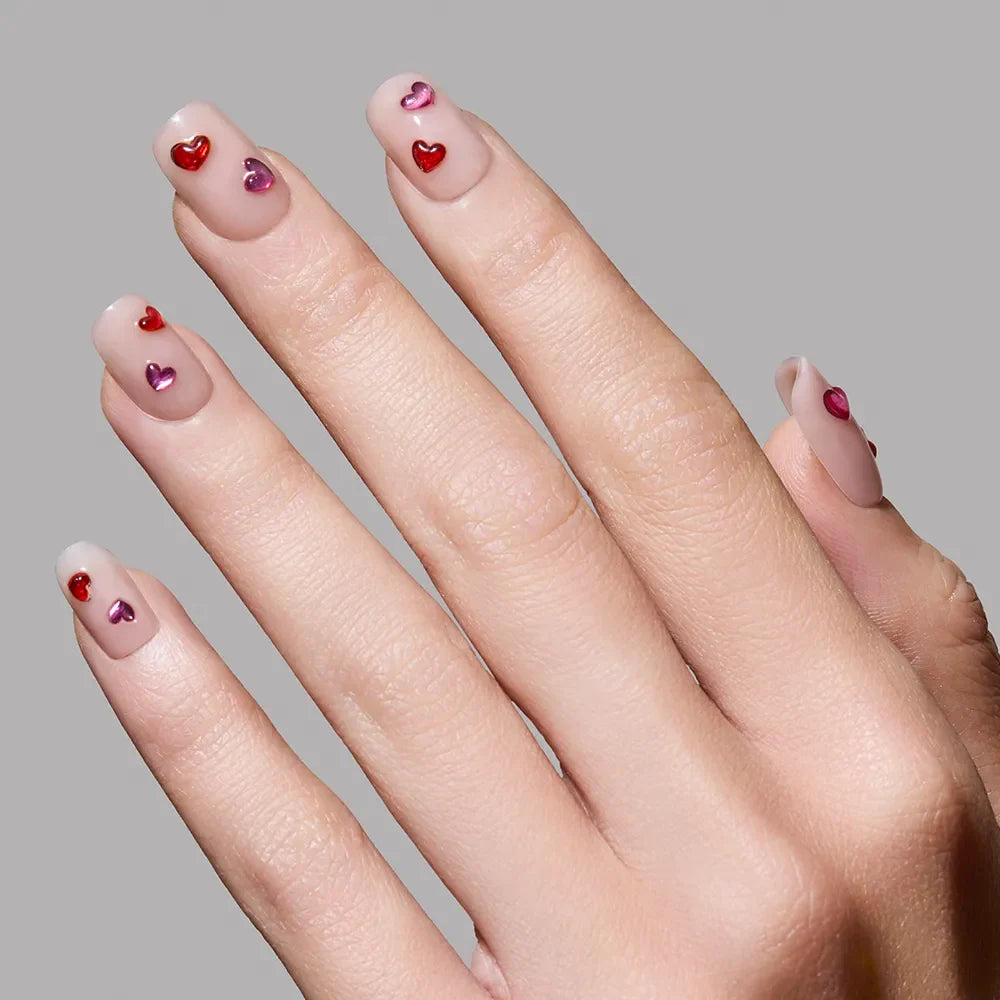 A hand showcases glamnetic’s Dear Lover nails: glossy nude, short squoval tips, each with shiny red and pink 3D gemstone hearts, set against a plain gray background.