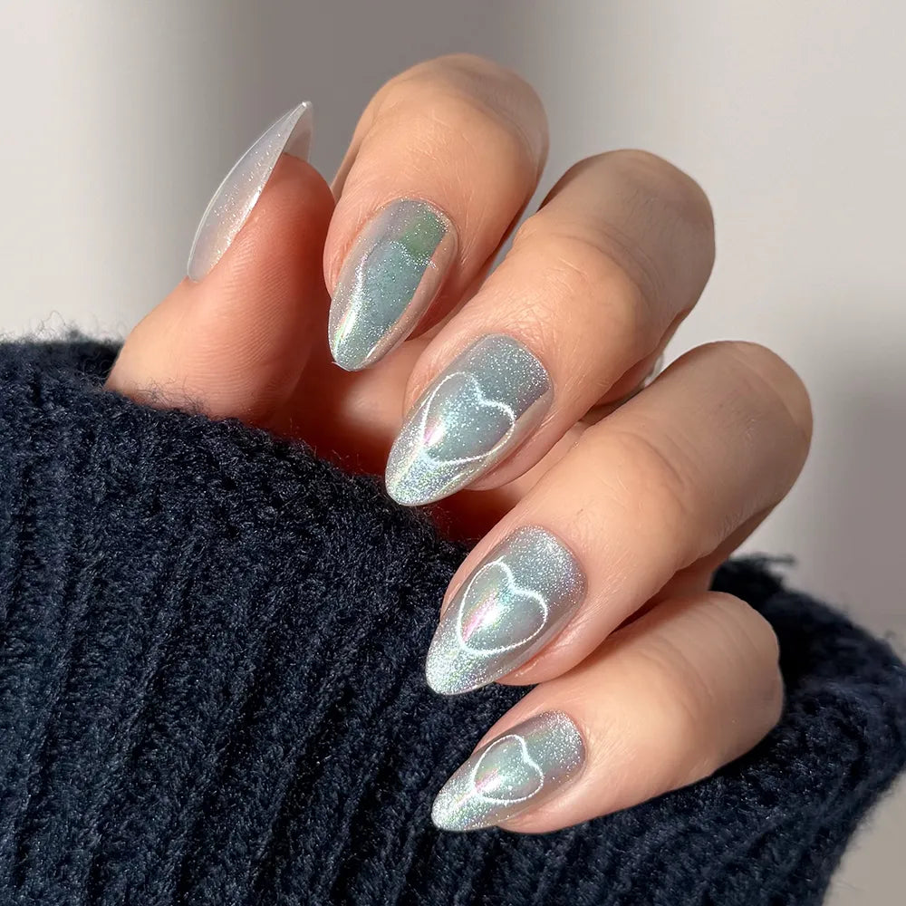 A hand with almond-shaped fairy-core nails painted in glamnetic’s Electric Lover, a shimmery iridescent light blue, each featuring a white heart outline. The person wears a dark, fuzzy sweater.
