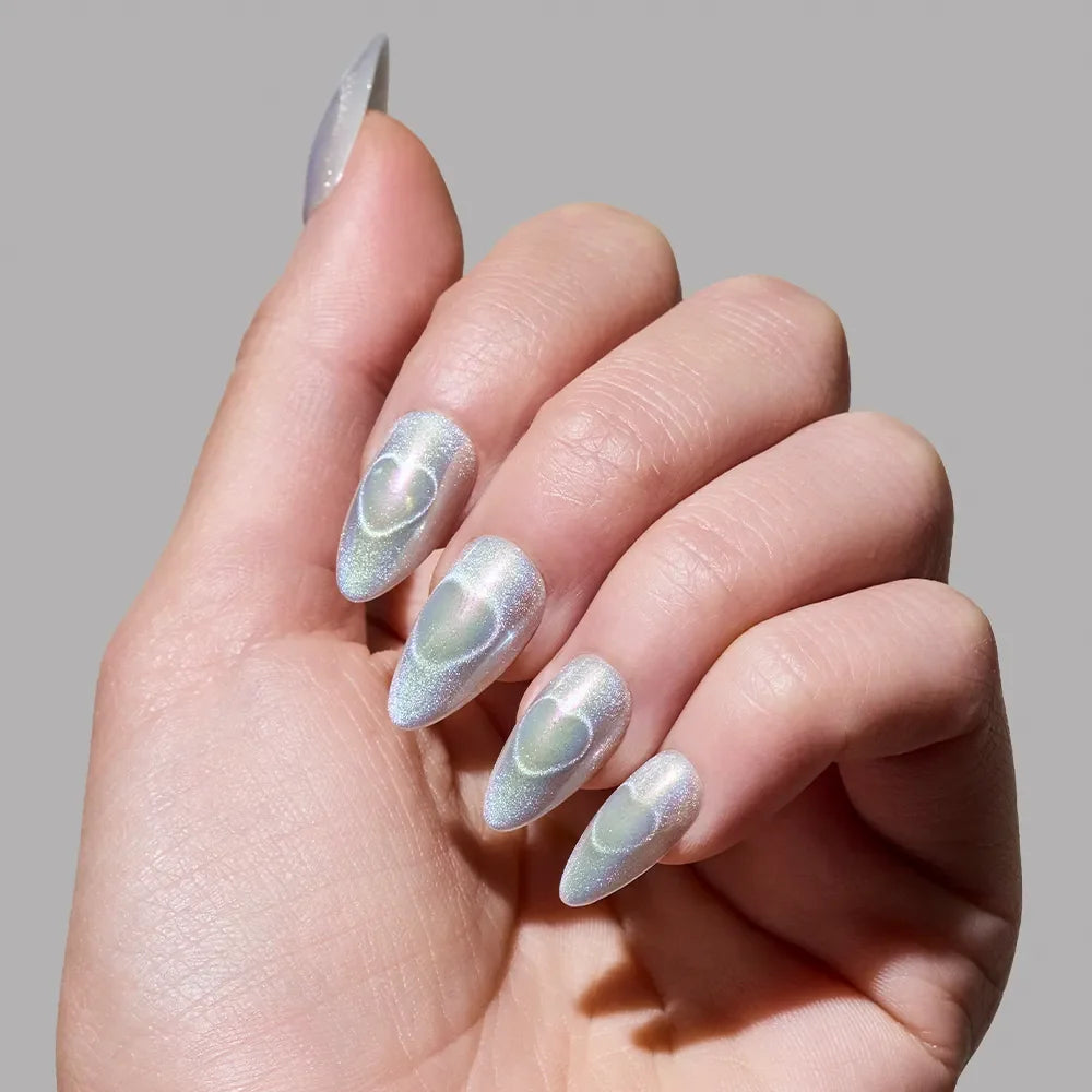A hand flaunts long, almond-shaped holographic nails by glamnetic in the shade Electric Lover, each with a subtle raised heart design atop shimmery iridescent polish, set against a plain gray background.