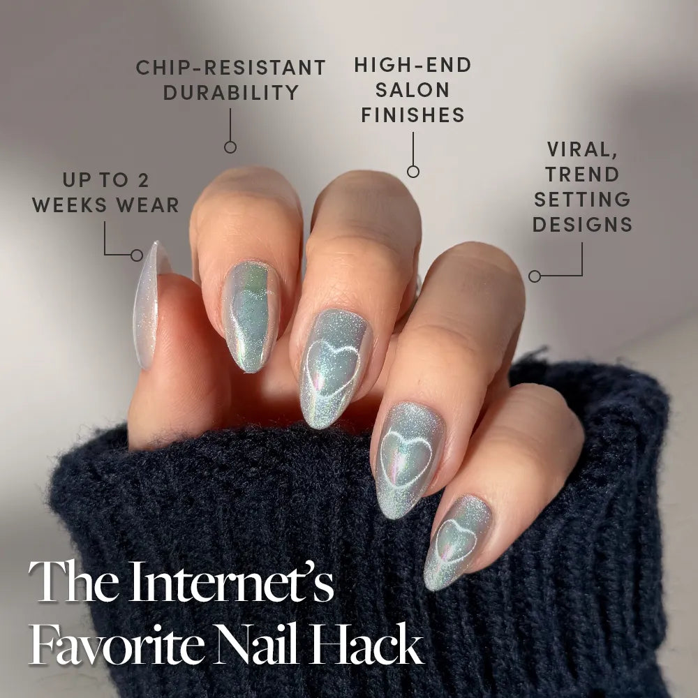 A hand with long, almond-shaped nails painted in glamnetic's Electric Lover—shimmery blue polish with white hearts for a fairy-core look. Features chip-resistant durability, high-end finish, viral design, up to 2-week wear. Sweater sleeves peek out.