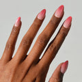 A hand with medium brown skin displays glamnetic Faded Feelings long almond press-on nails in a pink and white ombré against a plain light background.