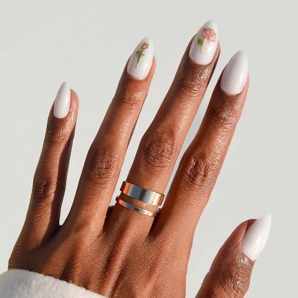 A hand with long, almond-shaped light pink nails from glamnetic's Floral Glow set—two adorned with 3D rose accents—models two gold rings against a plain light background.
