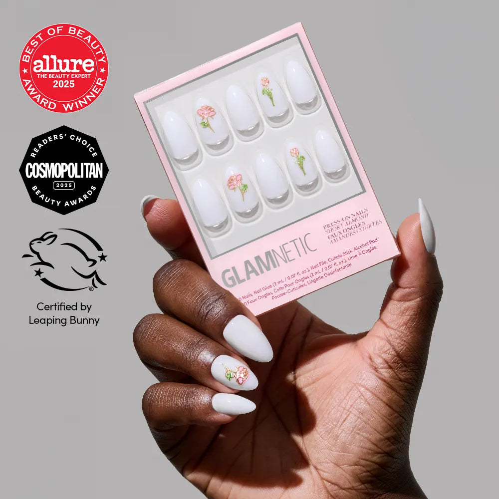 A hand with white short almond nails, some adorned with pink floral designs and 3D rose accents, holds a box of glamnetic’s Floral Glow nail set featuring Allure, Cosmopolitan, and Leaping Bunny cruelty-free award badges.