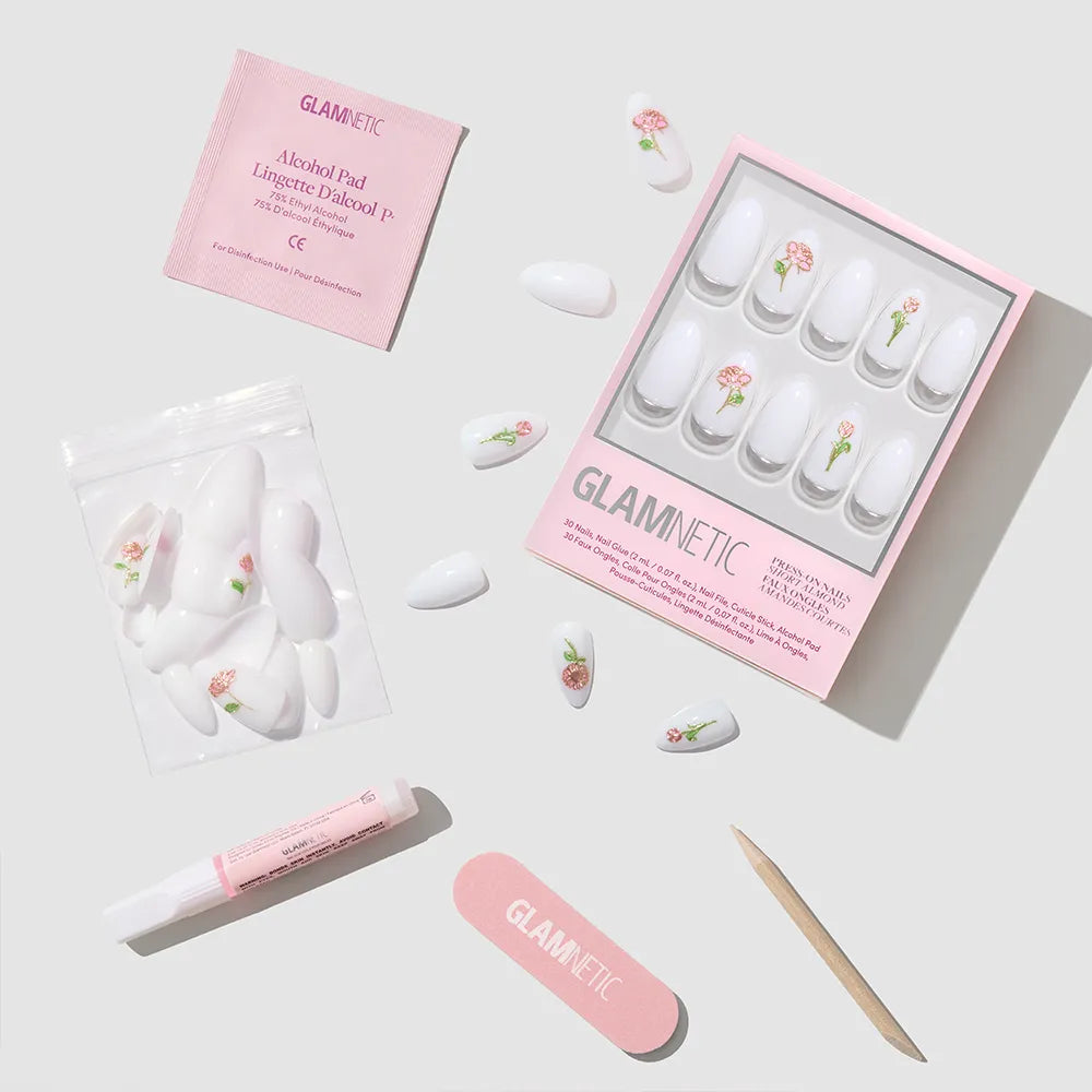 The glamnetic Floral Glow nail set features short almond nails with pink floral designs and 3D rose accents. It includes nail glue, a cuticle stick, a pink file, an alcohol pad, and extra nails displayed on a white surface.