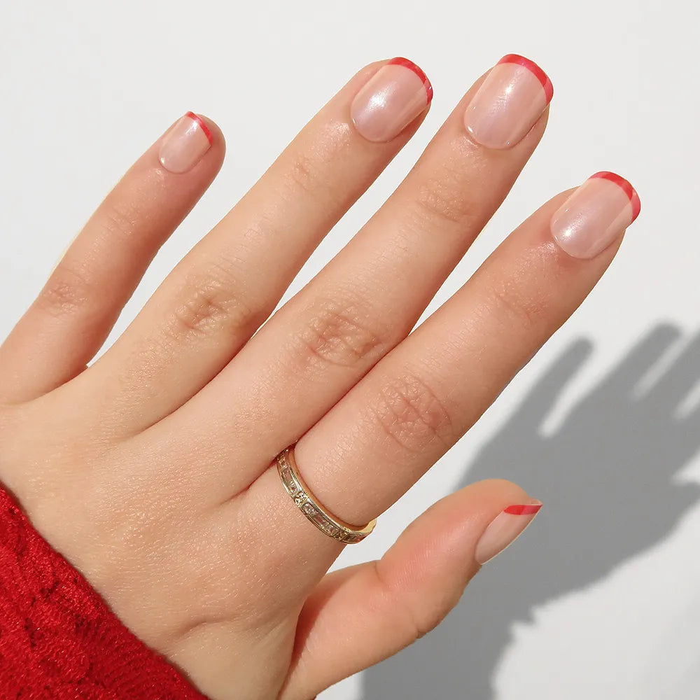 A hand wears a gold ring and sports a glamnetic Hotline Red micro French manicure on natural nails, paired with a red textured sleeve against a plain white background.