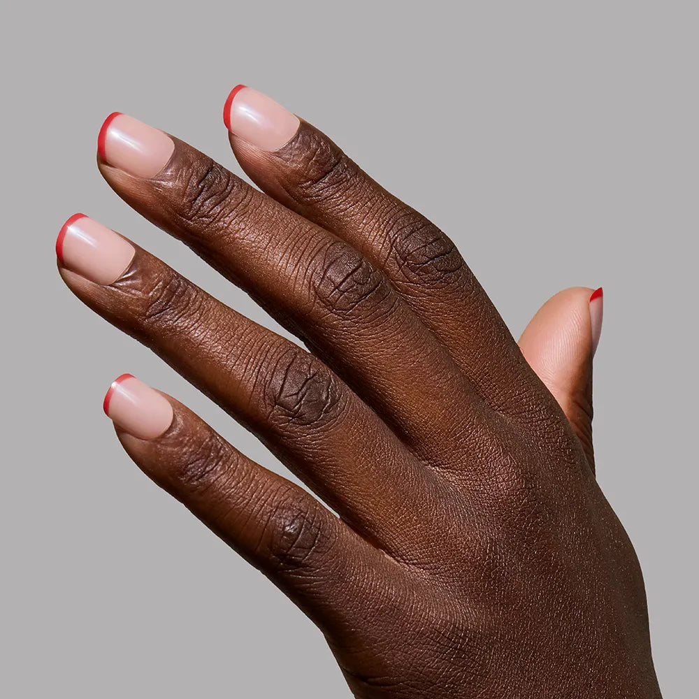 A hand with dark skin showcases neatly manicured nails with a nude base and red micro French tips using glamnetic's Hotline Red, set against a plain gray background.