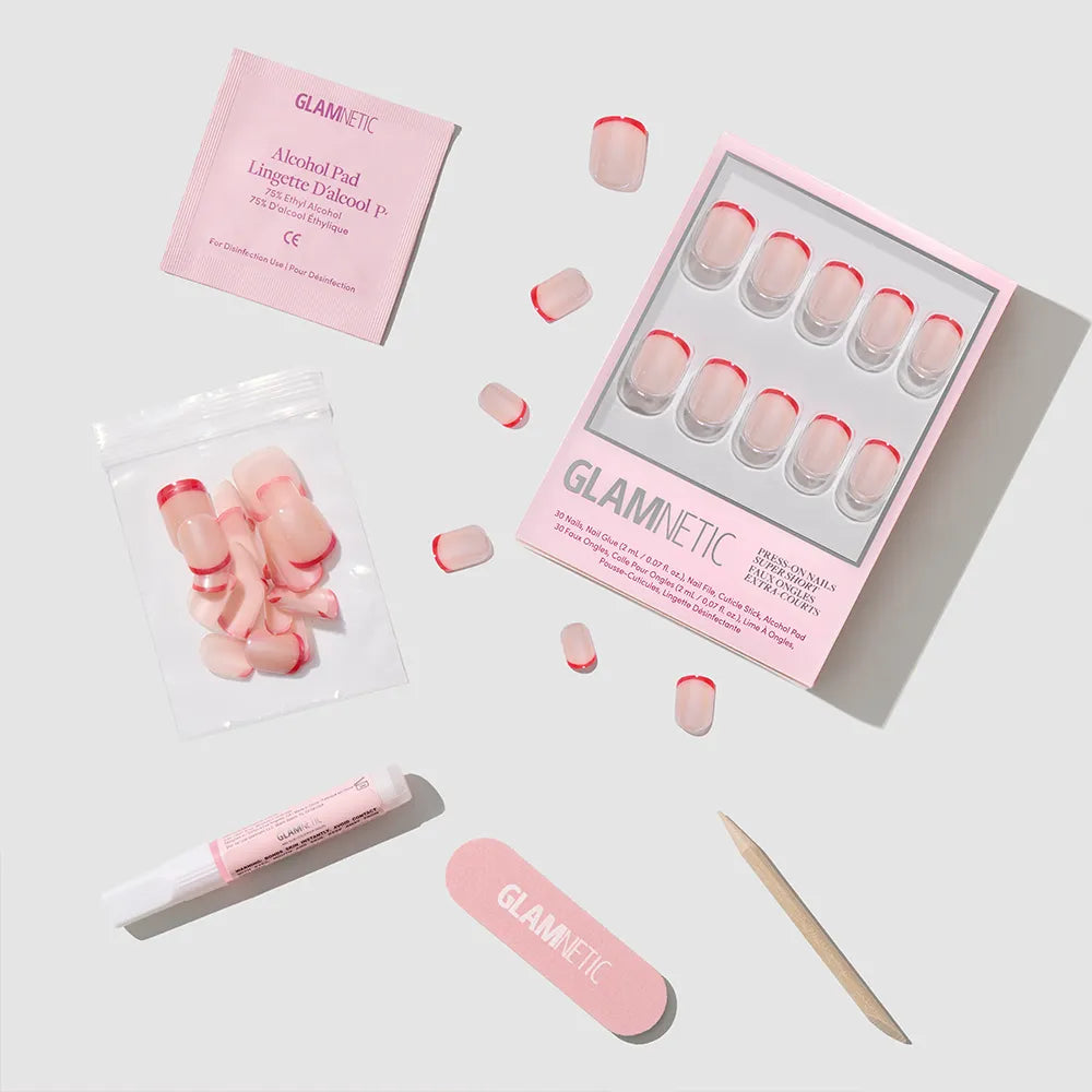 The glamnetic Hotline Red press-on nail set features pink nails with red micro French tips, nail glue, alcohol pad, nail file, cuticle stick, and extra nails—all arranged neatly on a light gray background.