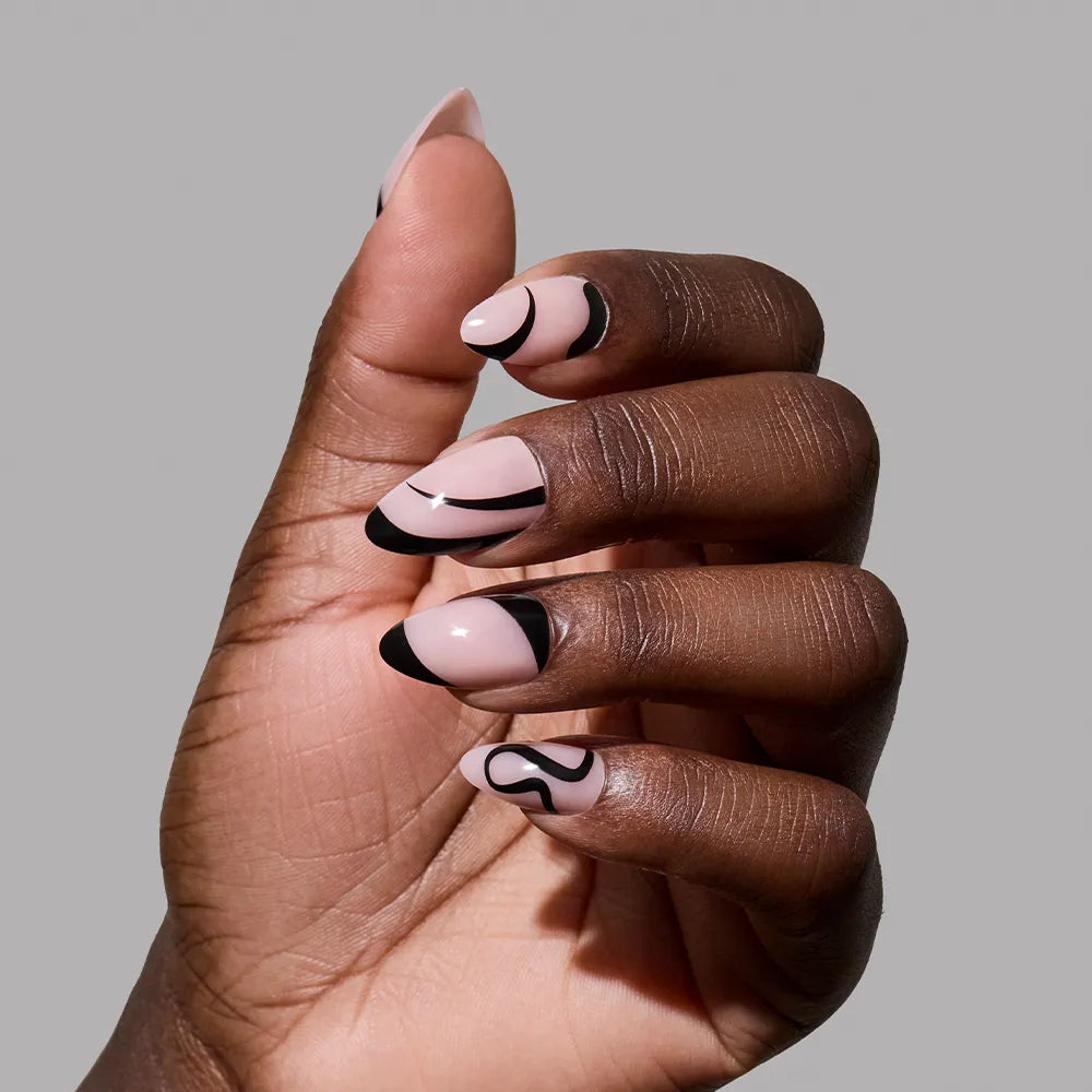 A hand with short almond nails wears glamnetic’s "Ink Wave" set—nude base nails detailed with black abstract lines and tips—highlighting a minimalist look against a plain grey background.
