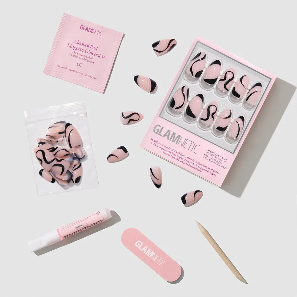 A flat lay of glamnetic’s Ink Wave press-on nail kit, featuring glossy pink and black swirl nails, nail glue, alcohol pad, nail file, wooden cuticle stick, and extra nails in a small bag.
