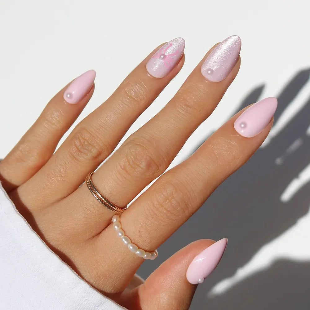 A hand wears glamnetic’s Love Knot long, almond-shaped pastel pink press-on nails with 3D pearl accents. Gold and pearl rings add to the soft-girl vibe, while a white sleeve contrasts the pristine background.