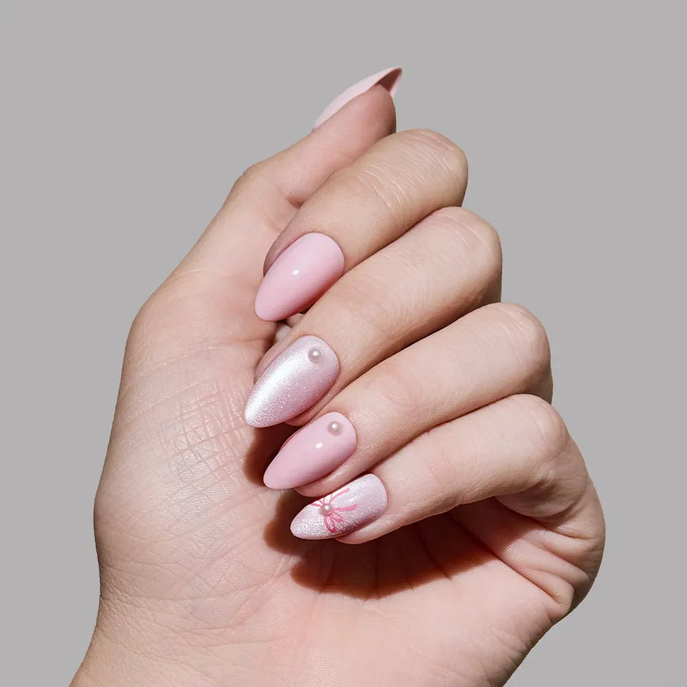A hand wears glamnetic Love Knot almond press-on nails in soft pink with shimmer, 3D pearl accents, and a delicate red lace design on one nail, set against a plain gray background.