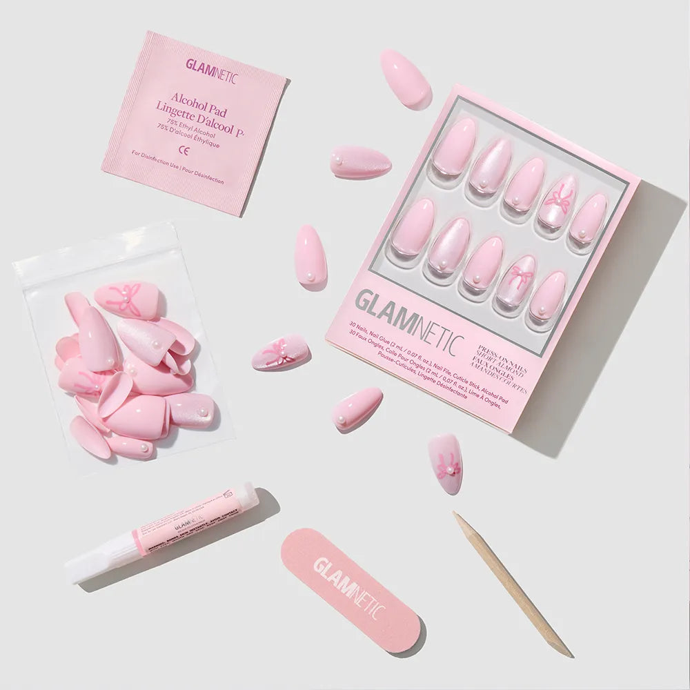 The glamnetic Love Knot set includes pink almond press-on nails with floral designs, a mini file, cuticle stick, adhesive tube, alcohol pad, and extra nails—all for the perfect soft-girl look on a light background.