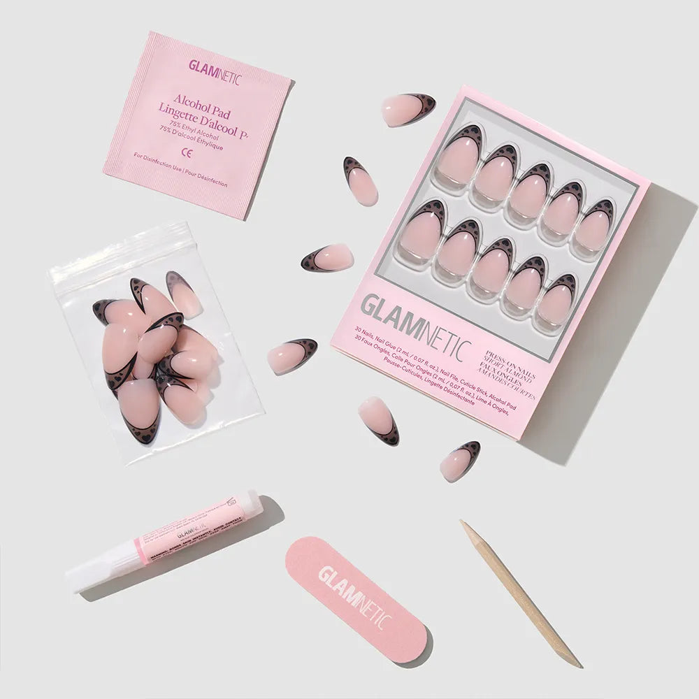 The glamnetic Love Noir set features pink and black press-on nails with a chic black heart design, a pink nail file, wooden cuticle stick, nail glue tube, alcohol pad, and extra nails—arranged neatly on a light gray surface.