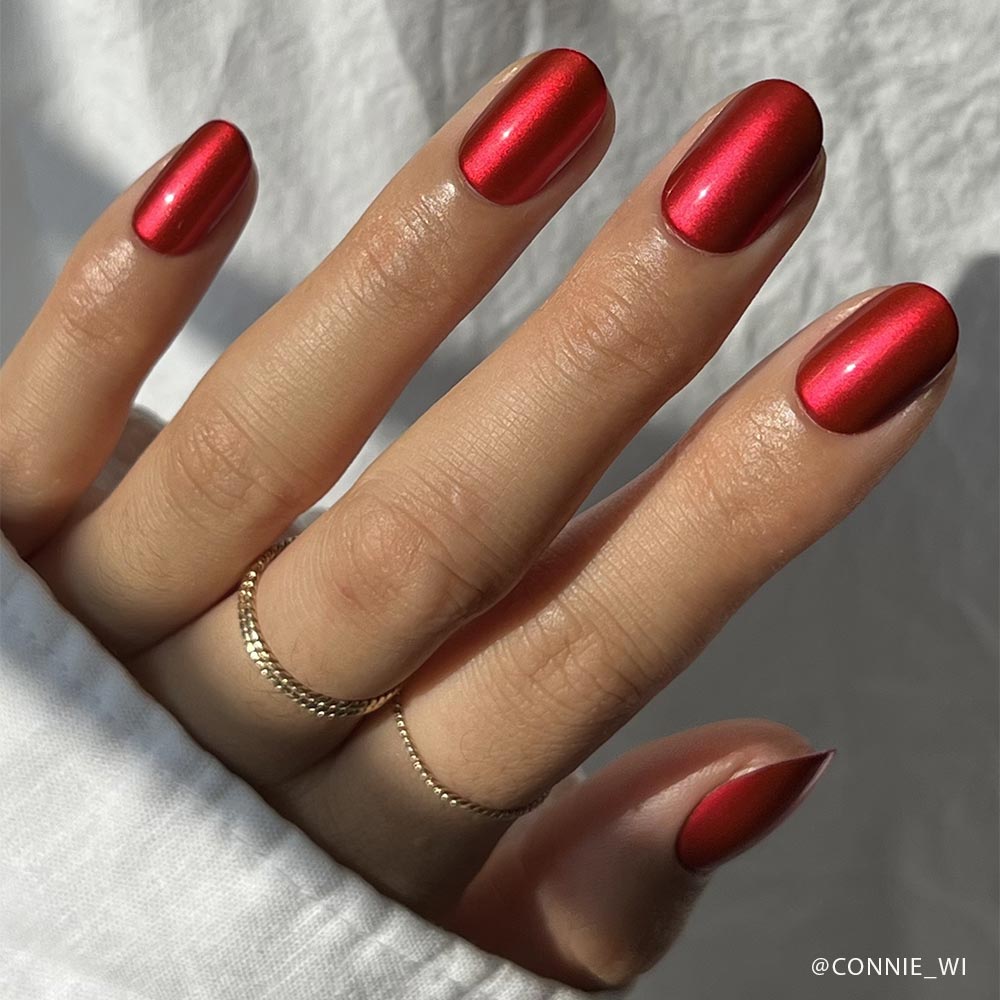 A hand with neatly manicured, shiny cherry-red nails in a short round style wears two gold rings and a white sleeve—showcasing the perfect look with glamnetics Cherry Glaze Nail Set.