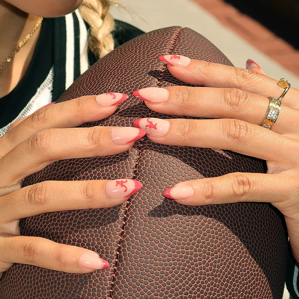 A person with long, almond-shaped glamnetic Alabama Crimson Tide Press-On Nails—pink with red tips and small red letters—holds a textured brown football. They wear a ring, black-and-white jacket, and have a blonde braid.