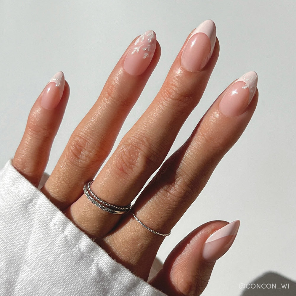 A hand with short almond nails in glamnetics All Time Love: a pink base, delicate French tips, and white floral nail art. The vegan, cruelty-free manicure is styled with two thin silver rings and a white sleeve against a plain background.