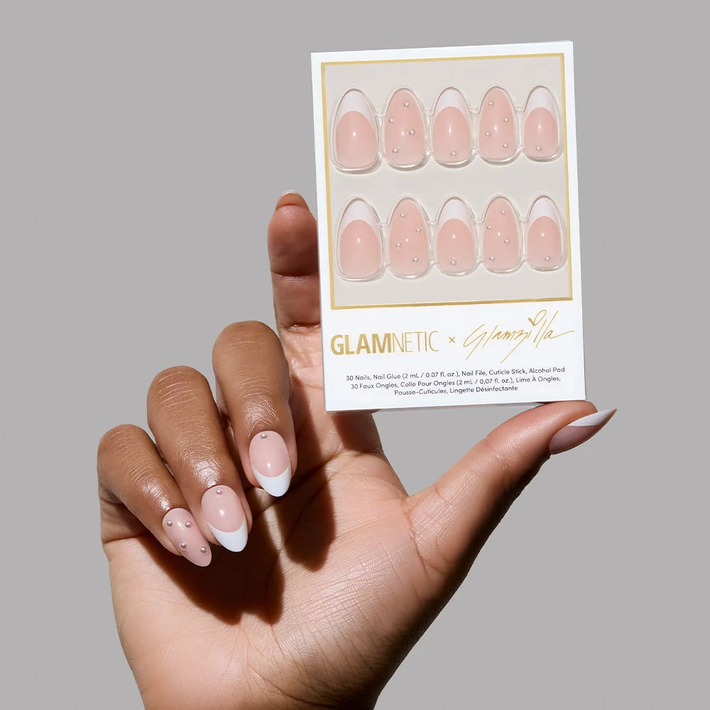 A hand holding the boxed Glamnetic x Glamzilla nail set, revealing 30 press-on nails in a pink base with a mix of white French tips and scattered pearl embellishments. The packaging is signed “Glamnetic x Glamzilla.”

