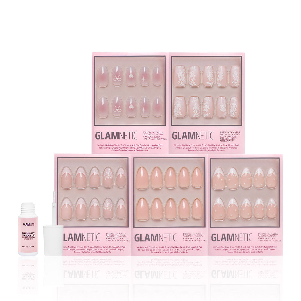 Six boxes of glamnetic Anns Sweethearts press-on nails in light pink and white, with floral, geometric, and French tip designs in a short almond style, are arranged in two rows. A small bottle of glamnetic nail glue is on the left.