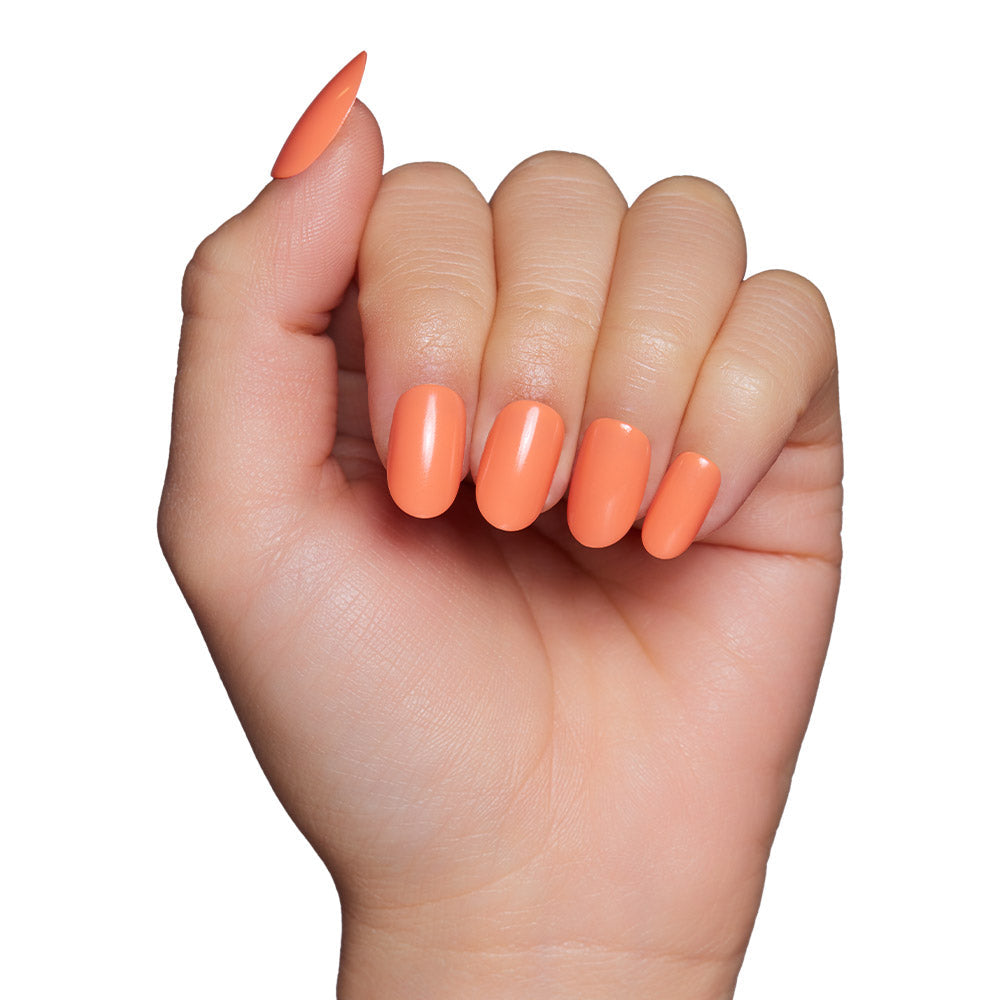 A hand with medium-length almond-shaped nails wears the glamnetic Apricot press-on set, shown in a glossy peach-orange polish against a white background.