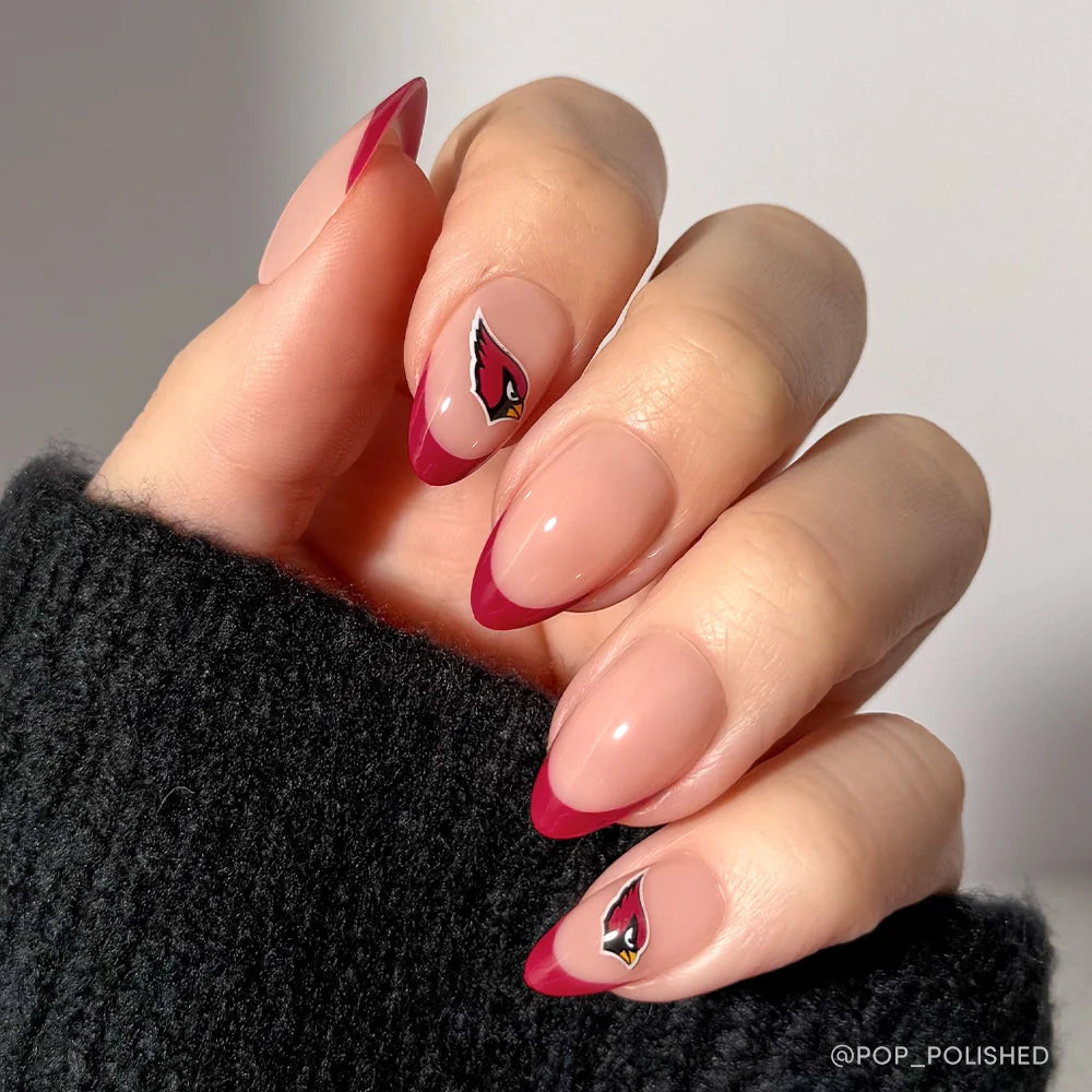 A hand with almond-shaped nude and dark red French tips displays the glamnetic Arizona Cardinals Press-On Nail Set, featuring team logos on the ring and pinky fingers. The person is wearing a black knit sweater.