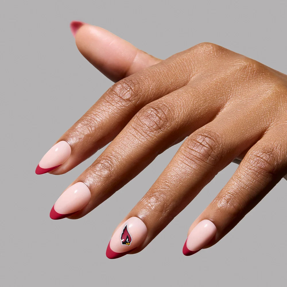 A hand showcases the glamnetic Arizona Cardinals Press-On Nail Set with almond French tips, a light pink base, red tips, and a cardinal bird on the ring fingernail, set against a plain gray background.