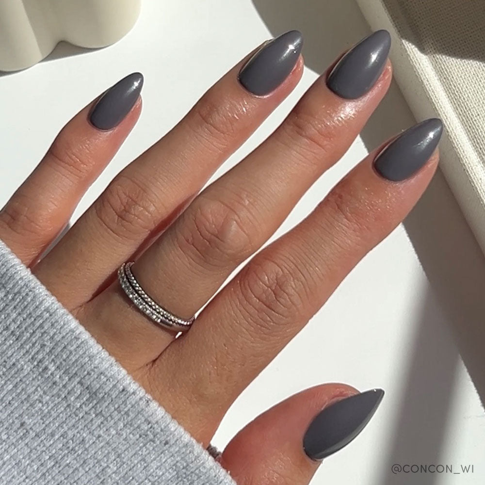 A hand with long, almond-shaped Ash Gray nails by glamnetic is polished in glossy grey. The person wears a silver double-band ring on the middle finger and a light grey long-sleeve top, with sunlight casting soft shadows.