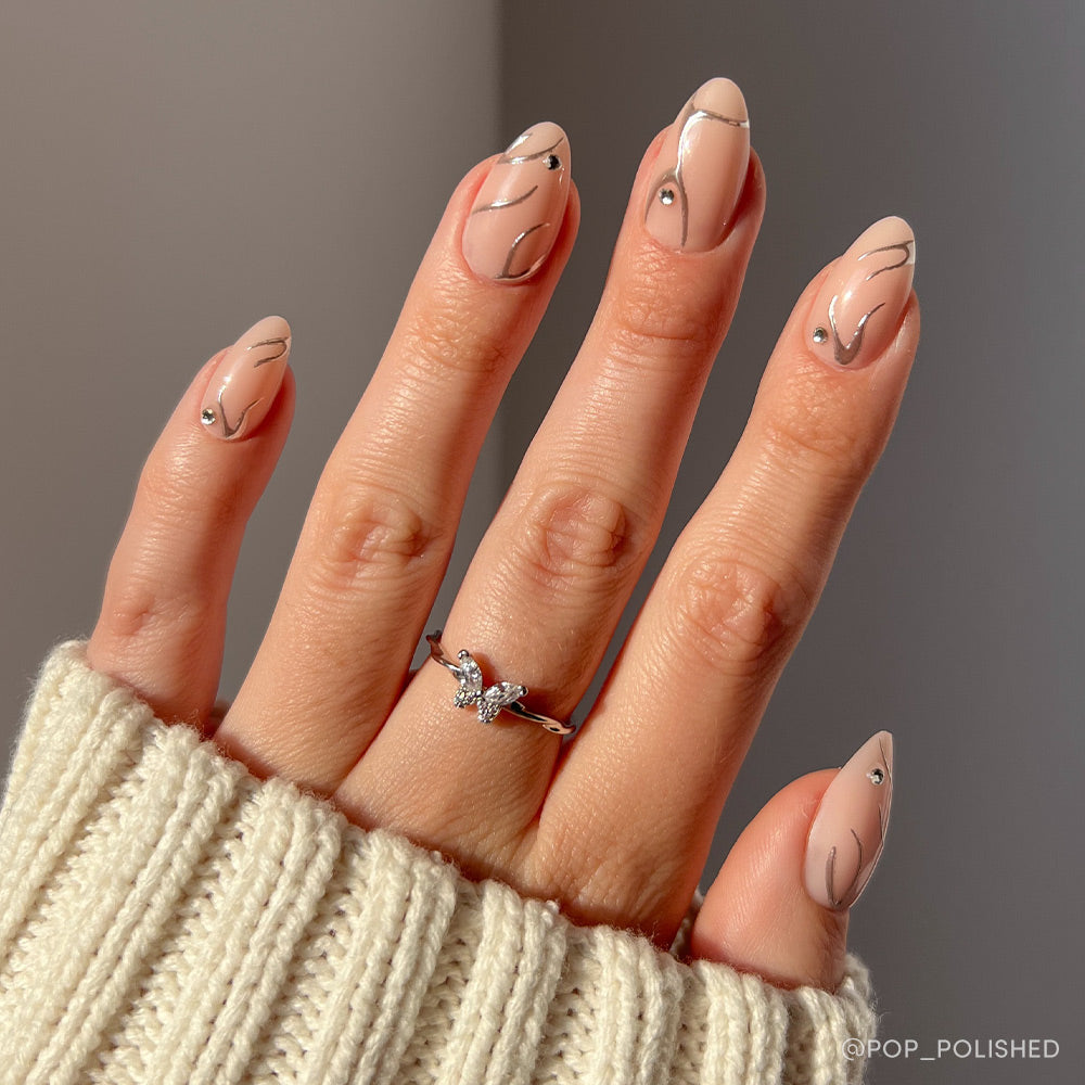 A hand with short almond-shaped nude Astro nails by glamnetic, featuring abstract black lines, rhinestones, and 3D chrome swirls; adorned with a silver butterfly ring and partially covered by a cream knit sweater.