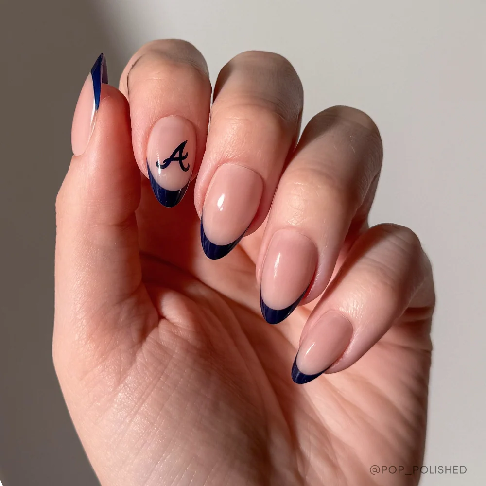 A hand with almond-shaped nails from glamnetics Atlanta Braves Press-On Nail Set: nude base, dark blue French tips, black letter A on the ring finger. Perfect for fans! Background is plain and light-colored.