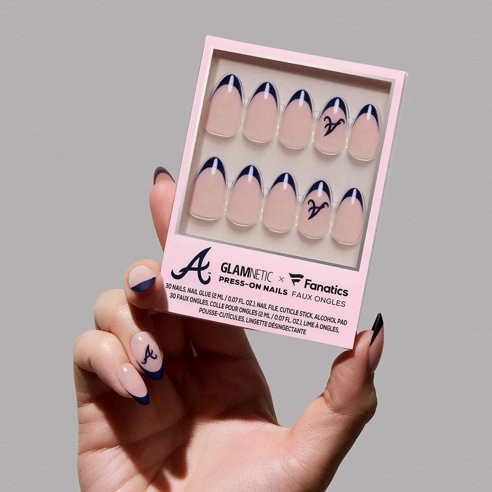 A hand with navy-tipped press-on nails holds a box of Glamnetic Atlanta Braves Press-On Nail Set, featuring a nude base, navy tips, and the iconic “A” logo for striking MLB-inspired nail art.