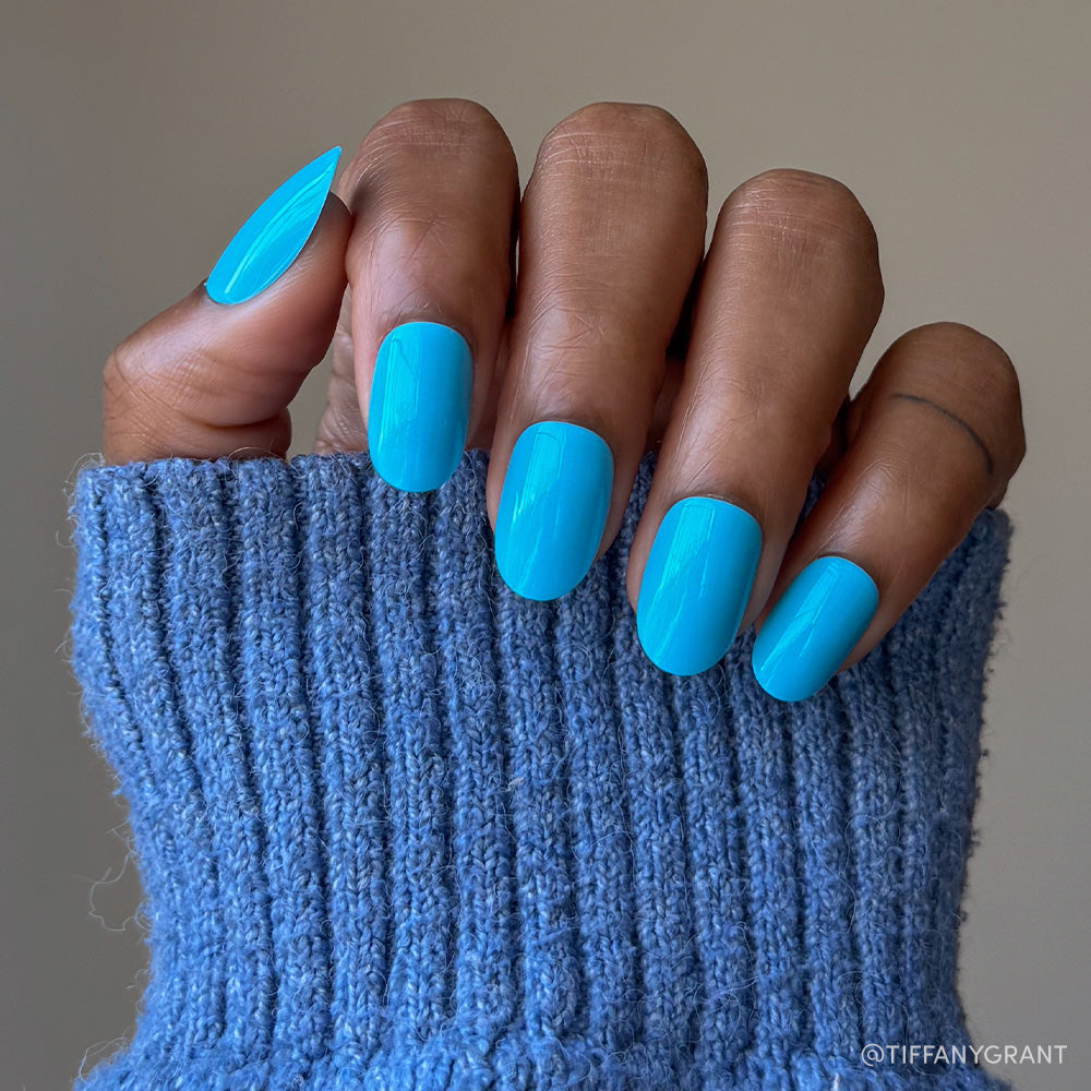 A hand with medium brown skin in a blue knit sweater displays neatly manicured nails painted with glamnetic’s Azure Blue, perfect for summer fun and long-lasting wear.