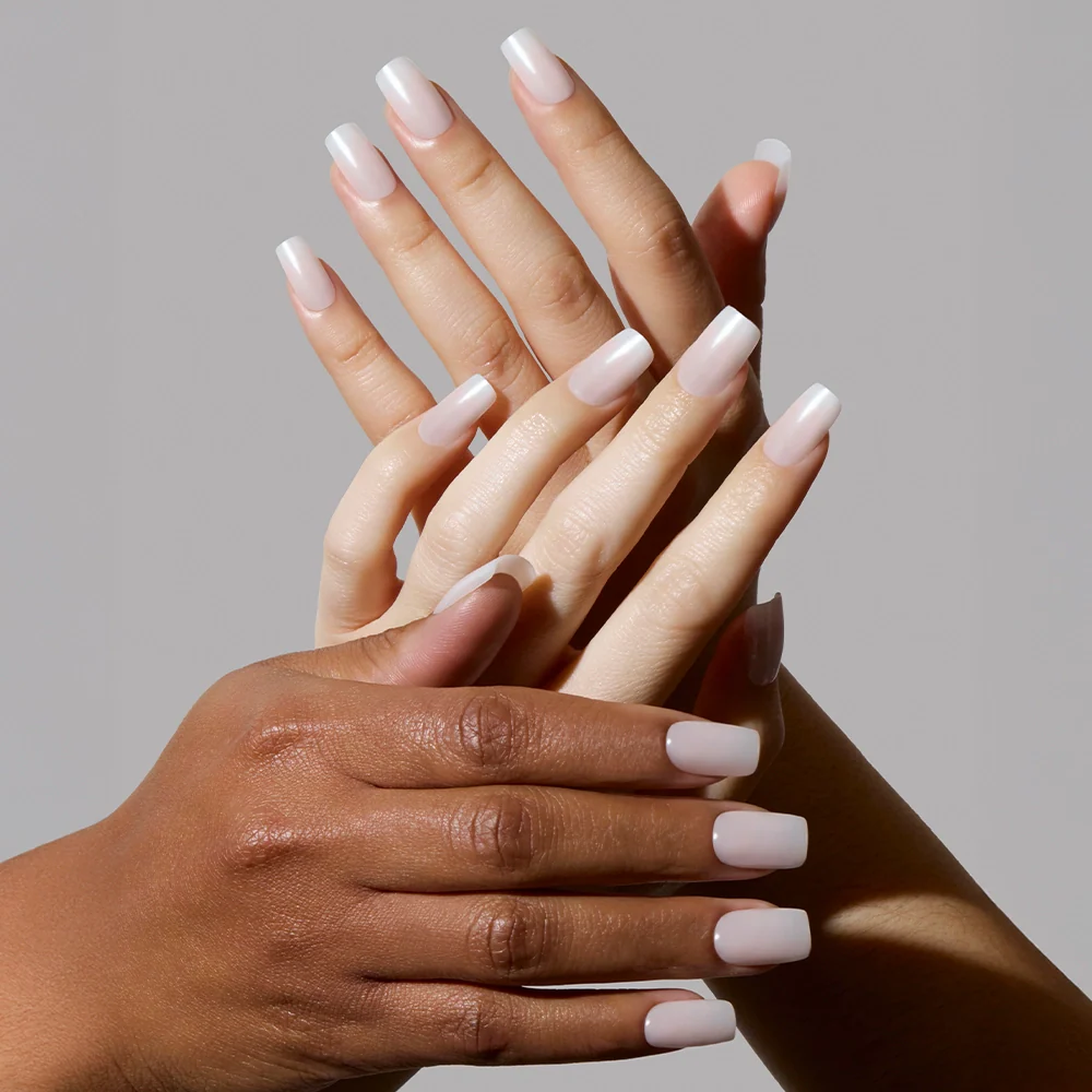 Two pairs of hands with different skin tones display Bailey medium square nails by glamnetic, featuring a glossy, semi-transparent light pink finish, posed gracefully against a plain gray background.