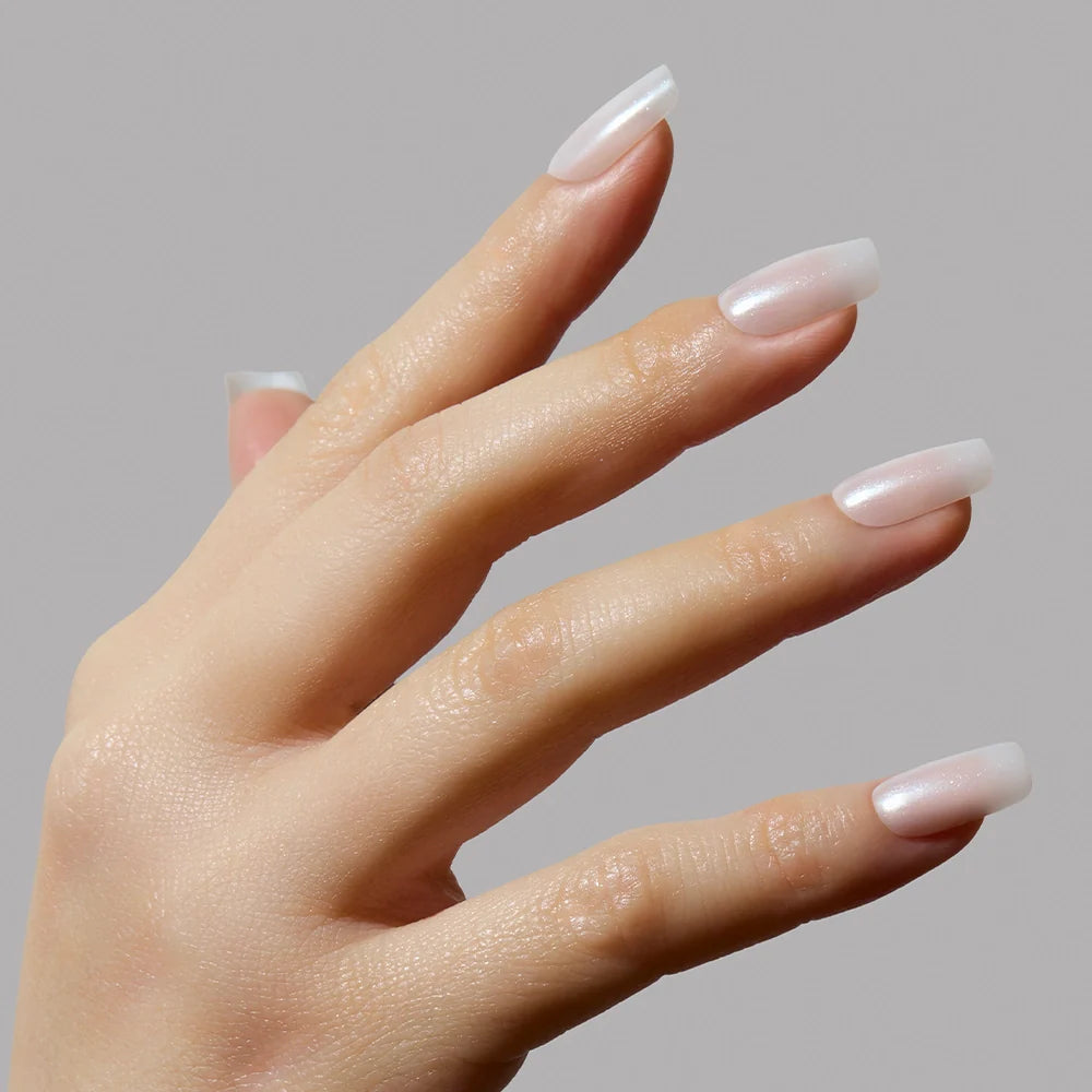 A hand with long, neatly manicured medium square nails painted in a glossy light pink Bailey shade by glamnetic is shown against a plain gray background.