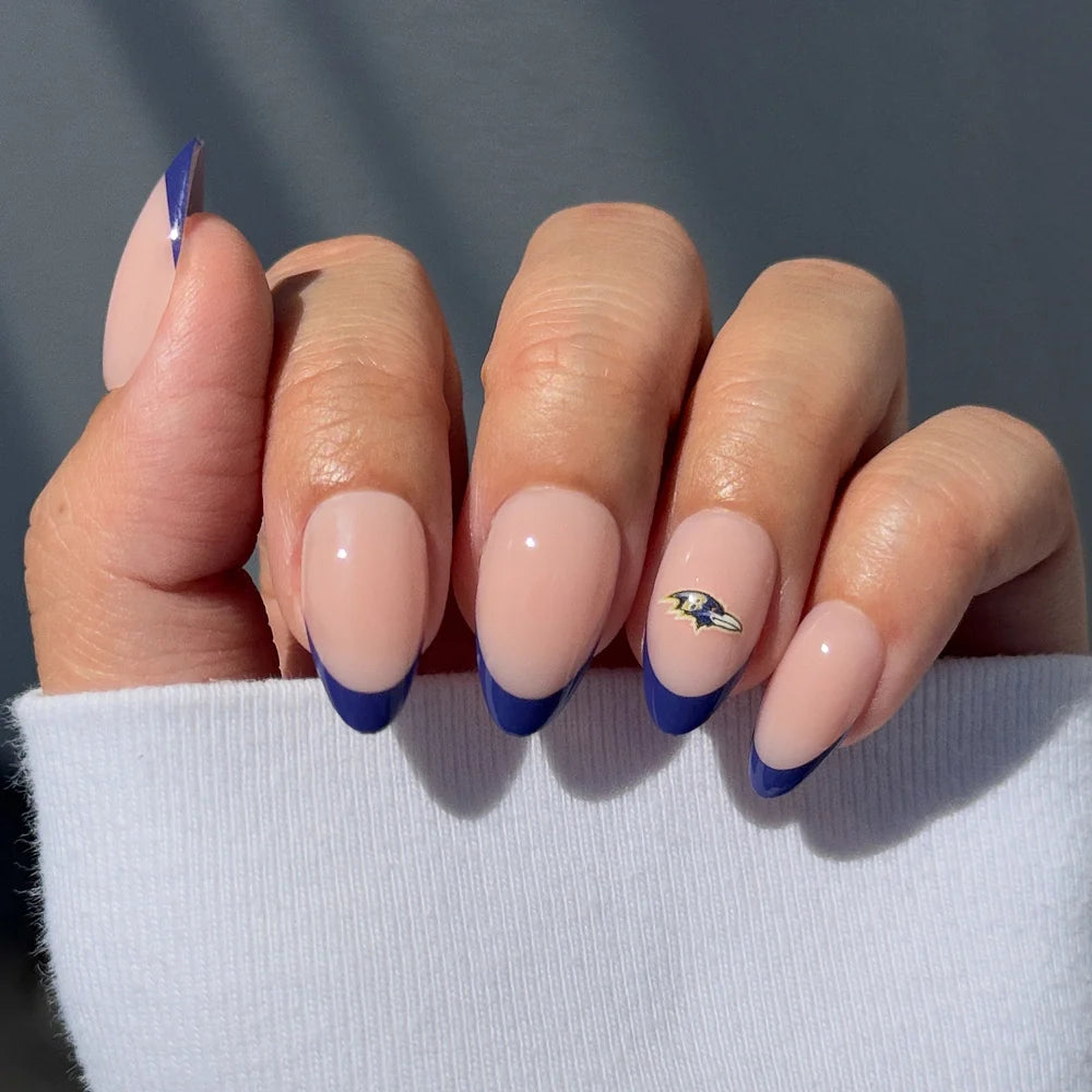 A hand with almond nails sports glamnetics Baltimore Ravens Press-On Nail Set—nude base, deep blue tips, and a metallic logo accent—making it perfect for football fans. The white sleeve partially covers the hand.