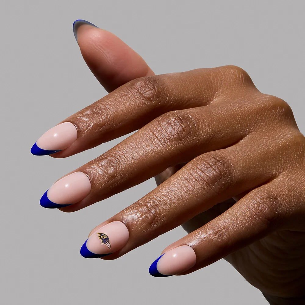 A hand with almond-shaped nails from the glamnetic Baltimore Ravens Press-On Nail Set—nude base, blue French tips, and a black-and-gold accent on the ring finger—perfect for subtle football fandom. The background is plain gray.