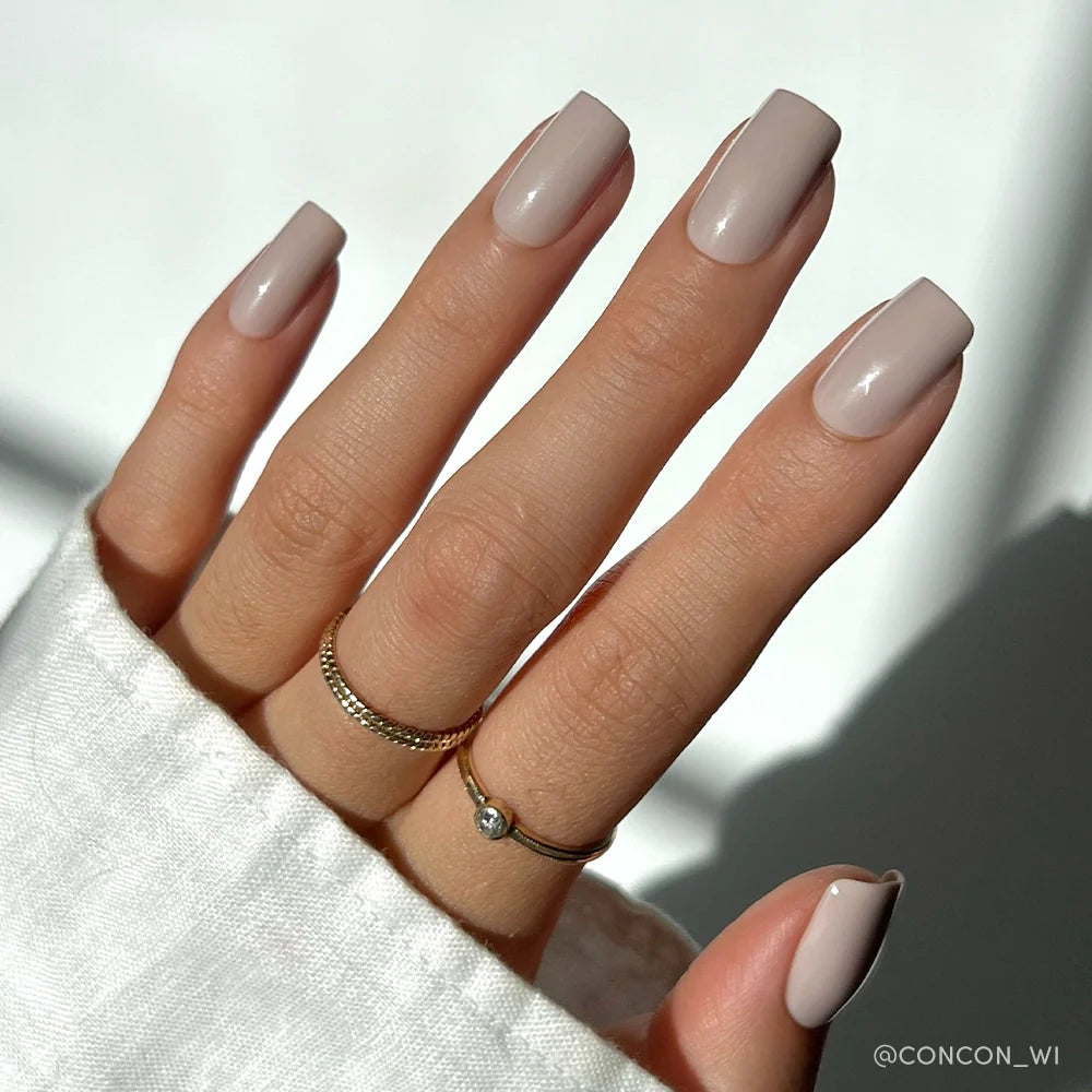 A hand with medium square nails, manicured in glamnetics Bare Bliss glossy nude set, exudes polished simplicity. Two gold rings and a white long-sleeve top complete the look against a softly lit background.