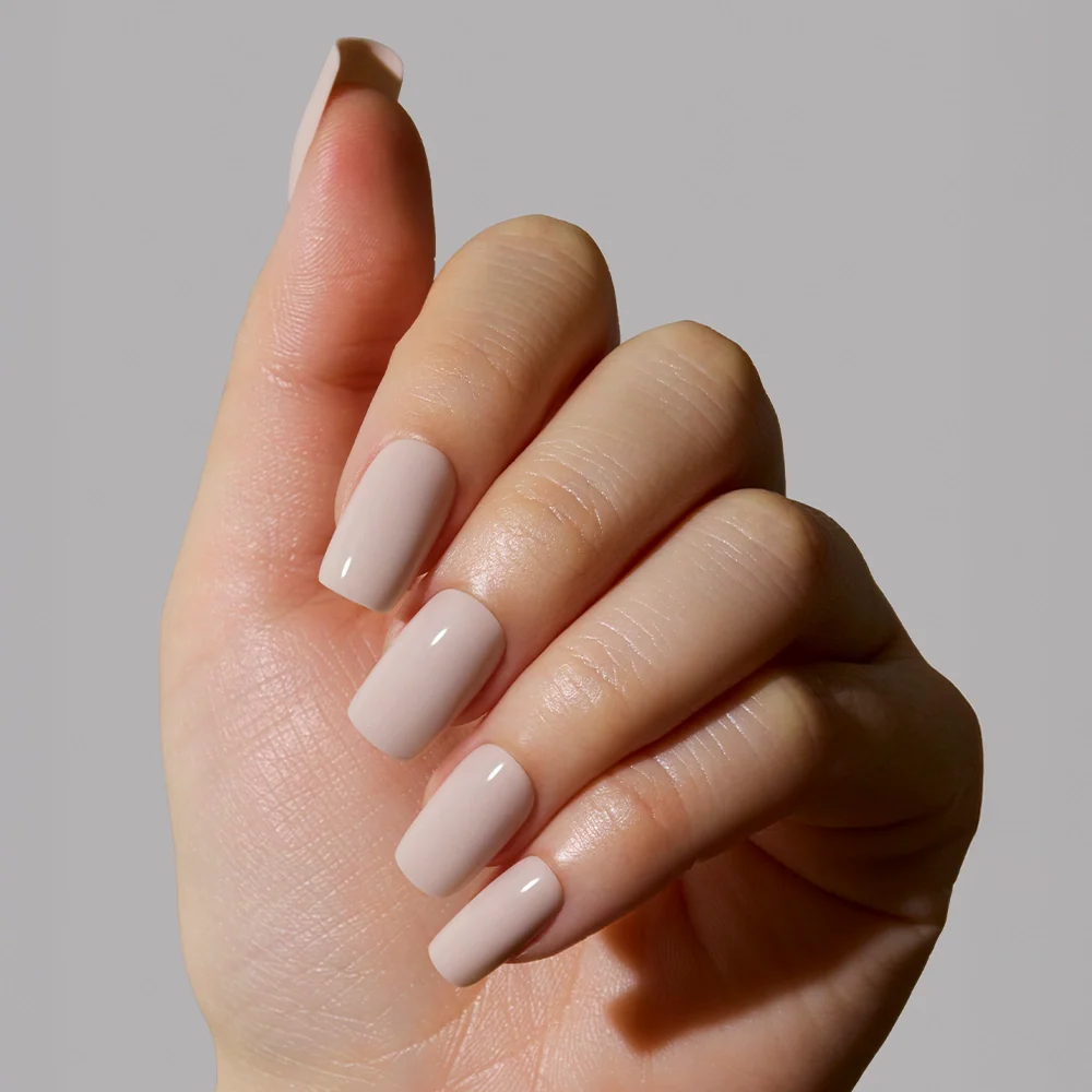 A hand with neatly manicured, medium square nails is shown against a plain light gray background. The nails are painted in glamnetics Bare Bliss, a glossy, opaque light nude shade for a polished look.